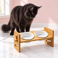 Kitty Buffet bamboo stand with two white ceramic bowls and a black Maine Coon looking toward the bowls in an empty light pink room with white marble flooring.
